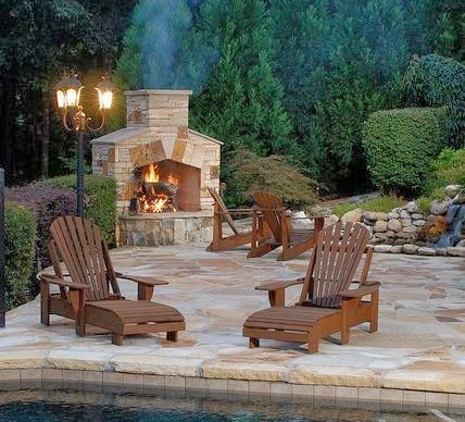 Outdoor fireplace with Adirondack chairs on a stone patio near a pool, surrounded by greenery.