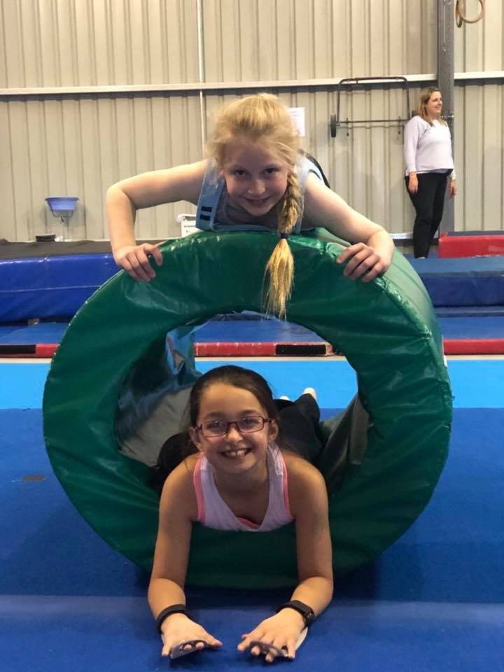Two young girls are playing with a green tire in a gym.
