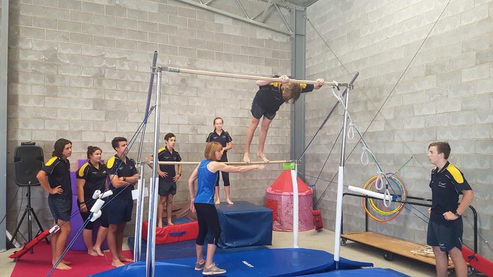 A group of people are watching a girl do a trick on a bar in a gym.