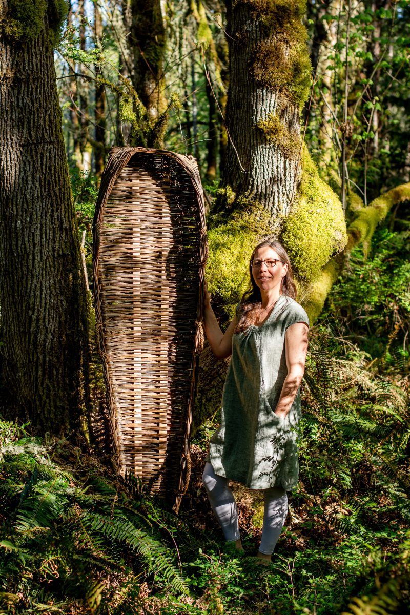 Photo of lush forest, old moss covered trees with a woman standing in the foreground with her hand on an upright willow casket
