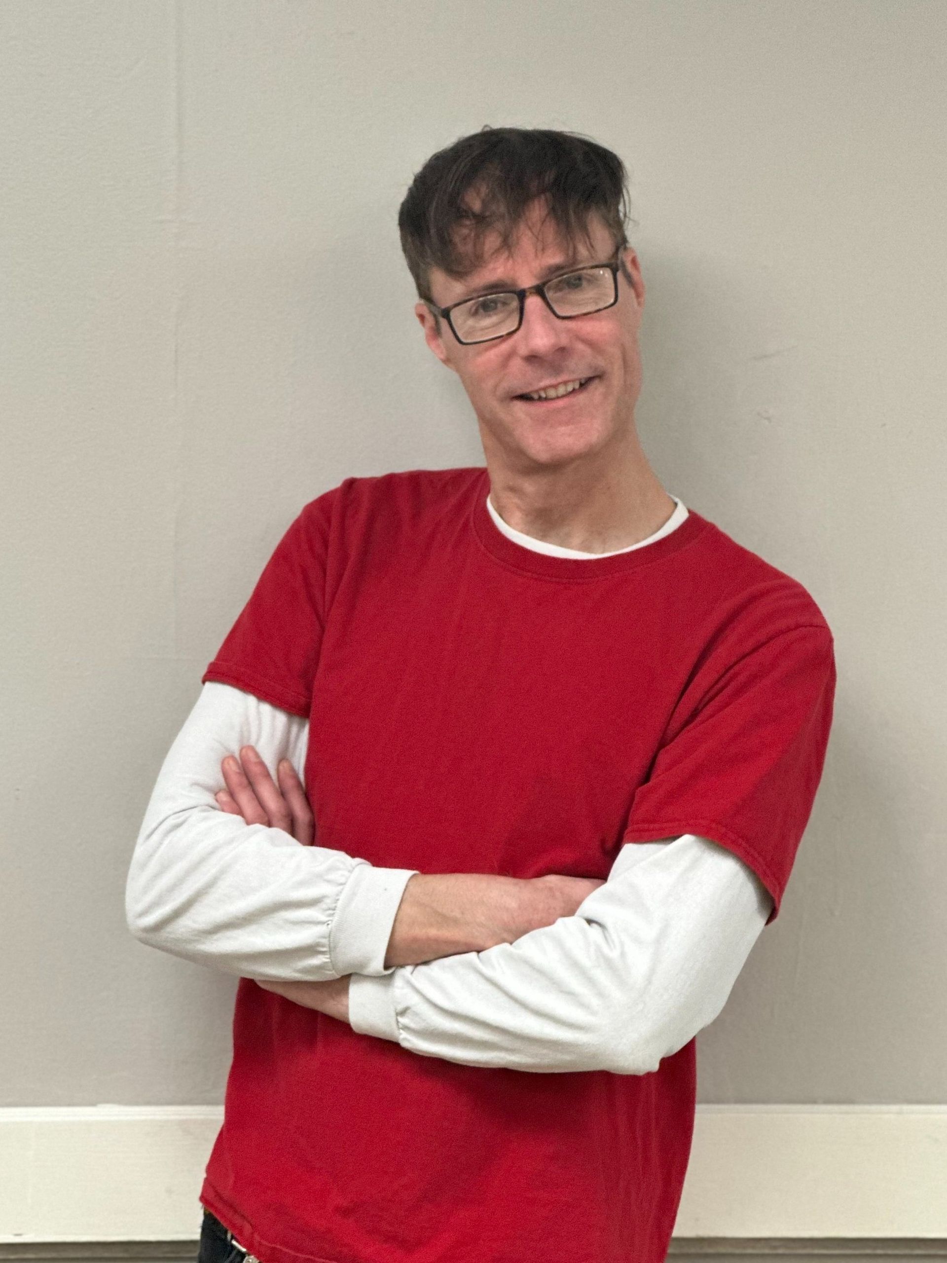 A man wearing glasses and a red shirt is standing with his arms crossed