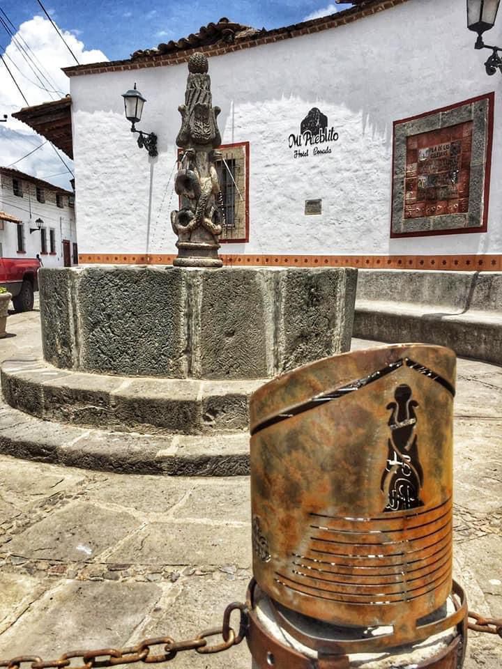Una fuente de piedra con un pilar tallado  en una plaza empedrada frente a un edificio blanco con un mural ornamentado.