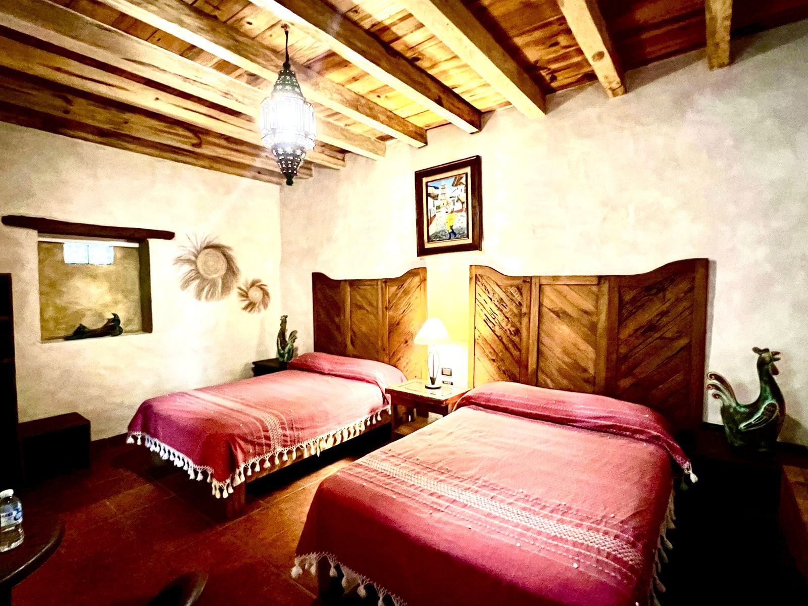 Habitación rústica con dos camas, cabeceros de madera, vigas de techo a la vista y una lámpara colgante estampada.