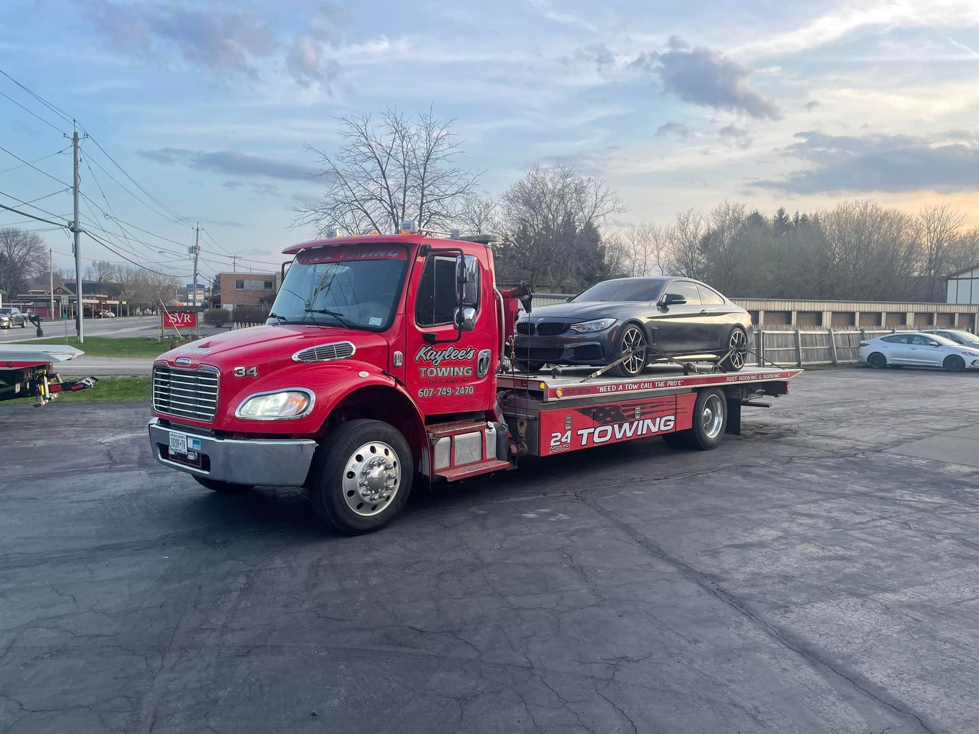 A red tow truck is towing a black car in a parking lot.