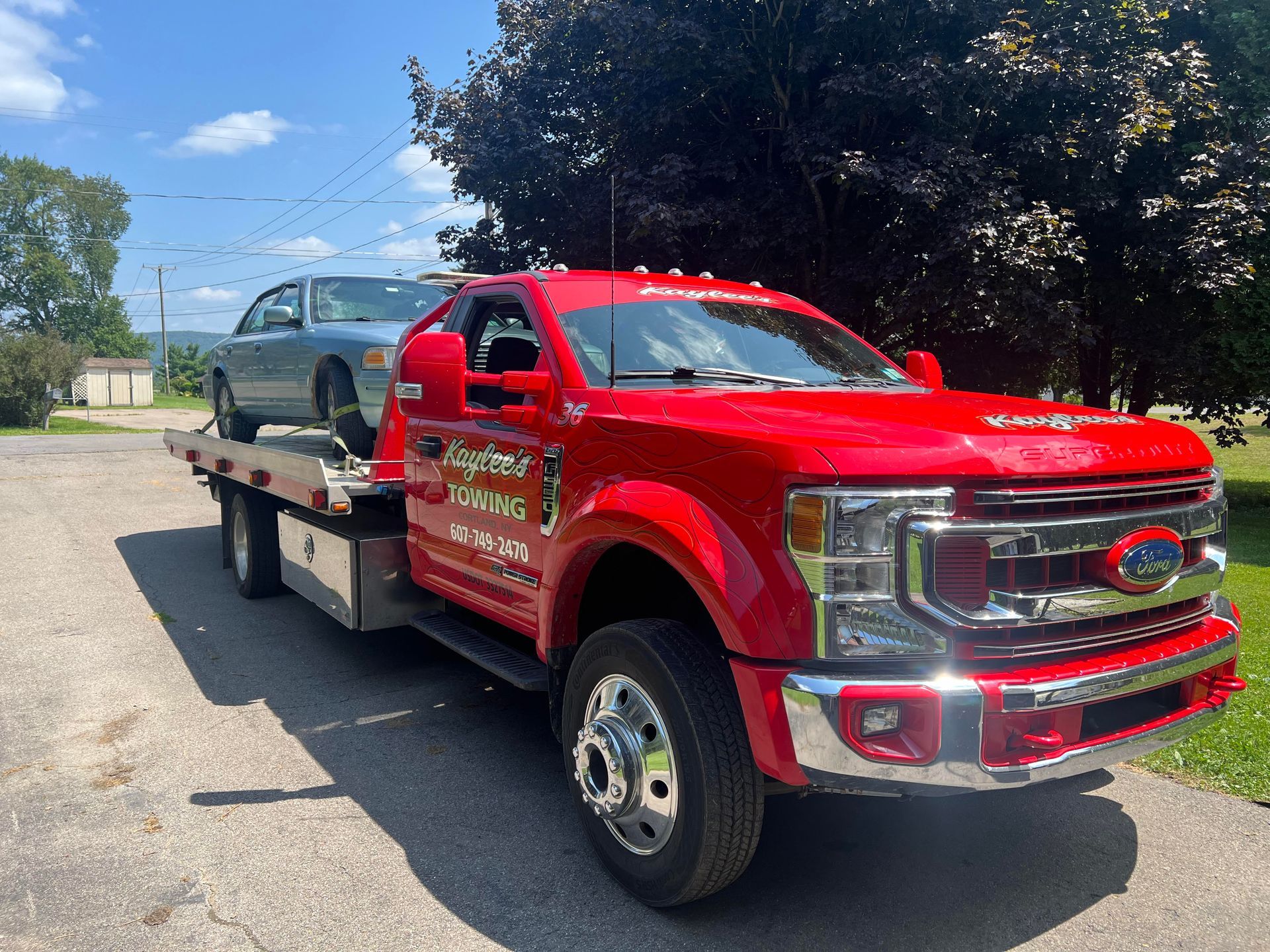 Red tow truck hauling a silver car on a sunny day. Truck has company name and phone number on the side.