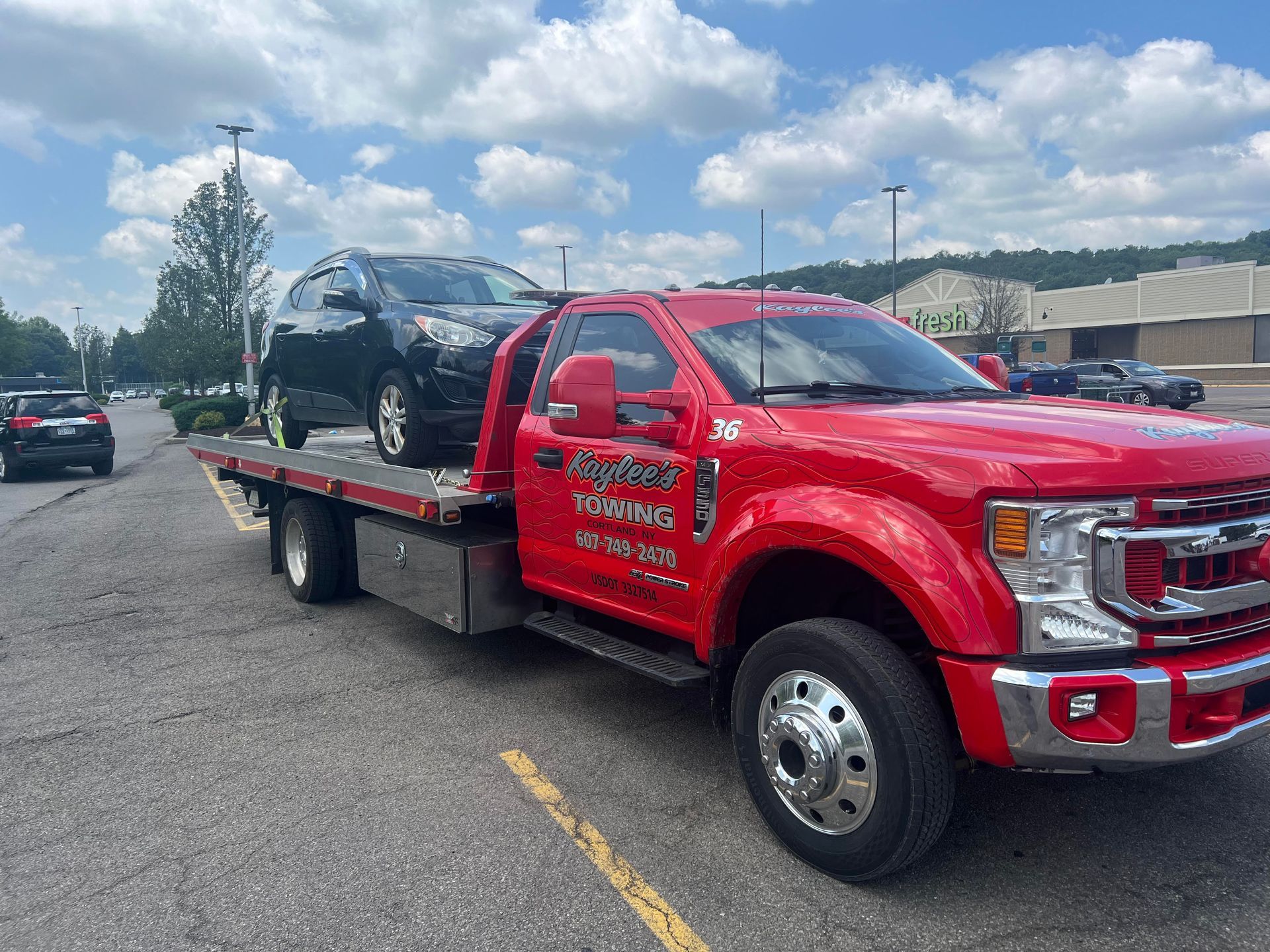 Red tow truck carrying a dark car in a parking lot on a sunny day. The truck has business information on the door.