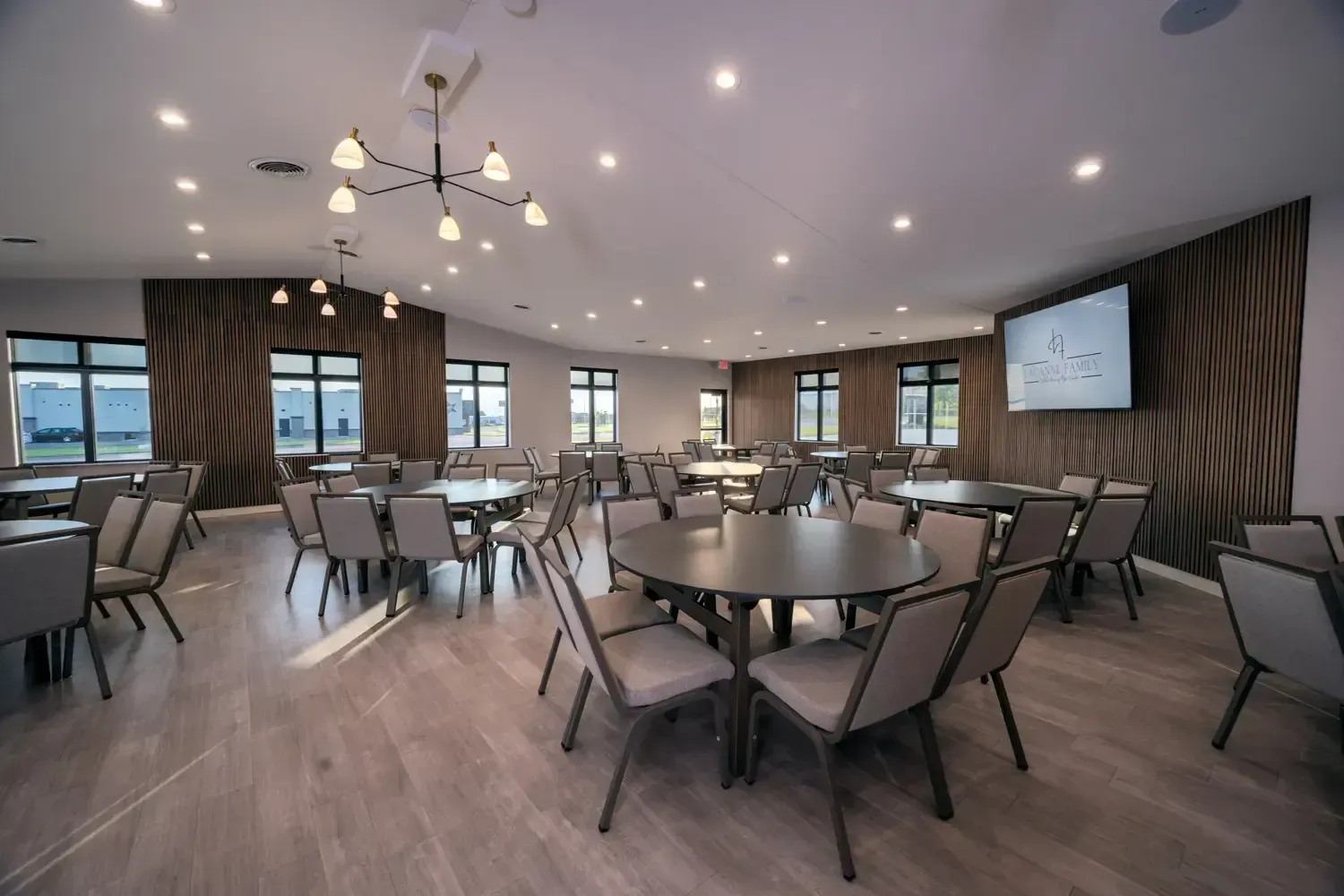 Spacious modern banquet room with round tables, chairs, chandeliers, and a wall-mounted screen.