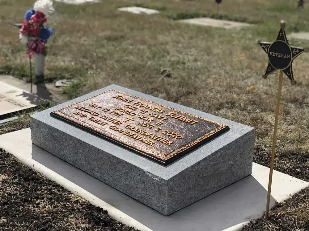 Gray memorial plaque on a concrete base in a grassy cemetery, with a star-shaped veterans marker beside it