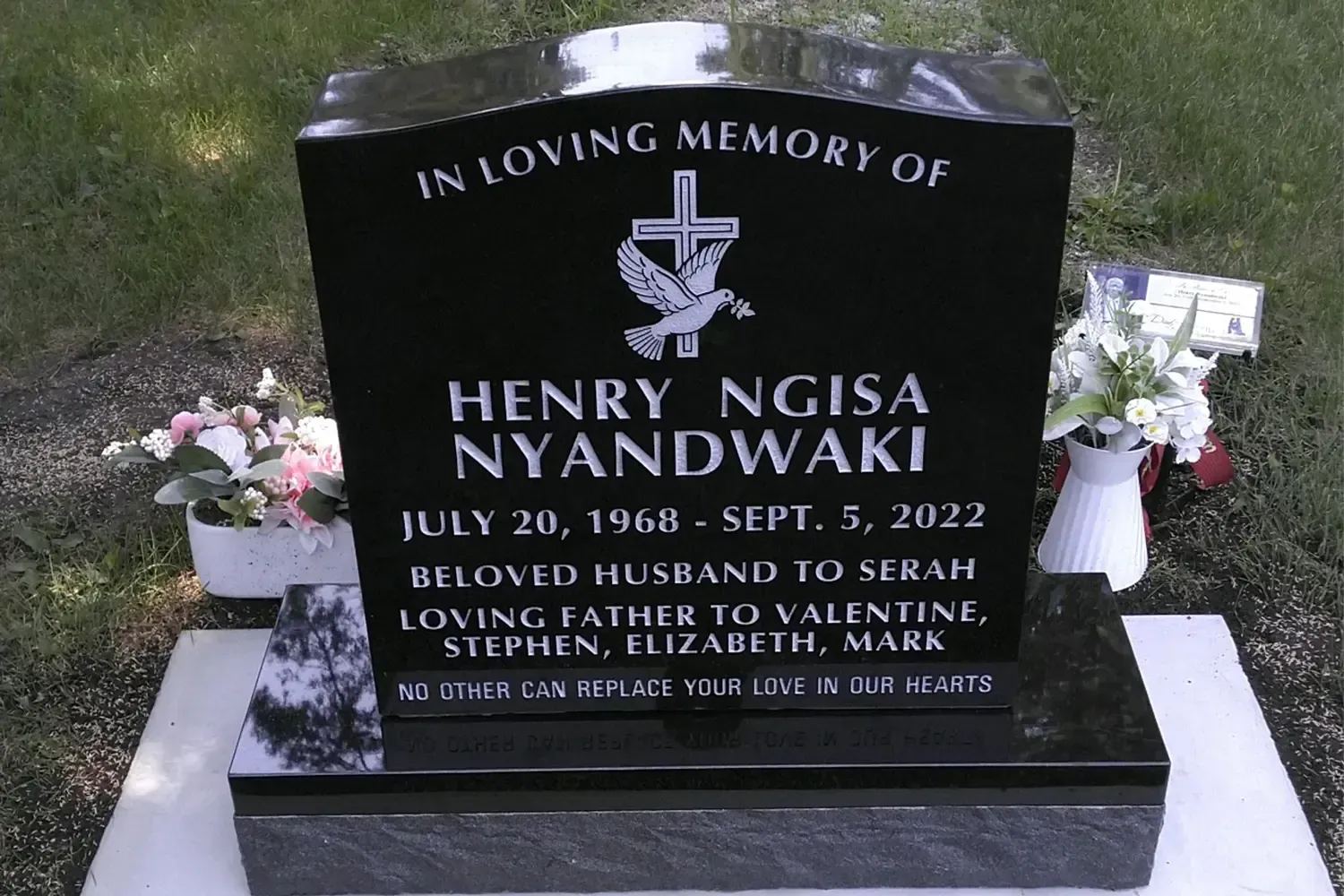 Black gravestone memorial for Henry Ngisa Nyandwahir, with flowers on a white pedestal outdoors.
