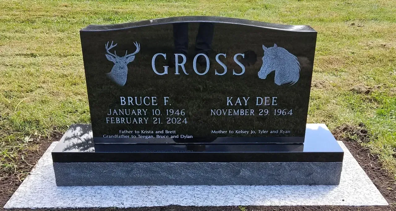 Black granite gravestone with deer and bear carvings, set on a concrete base in a grassy cemetery.