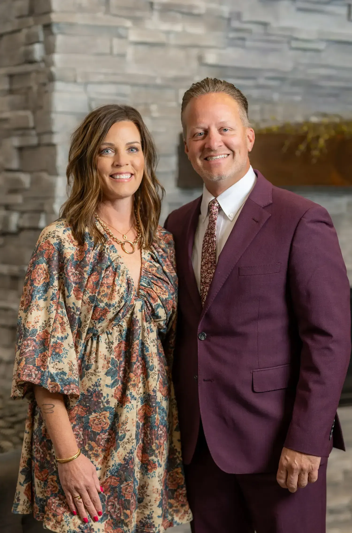 Two people standing together indoors, smiling in formal attire against a stone wall backdrop