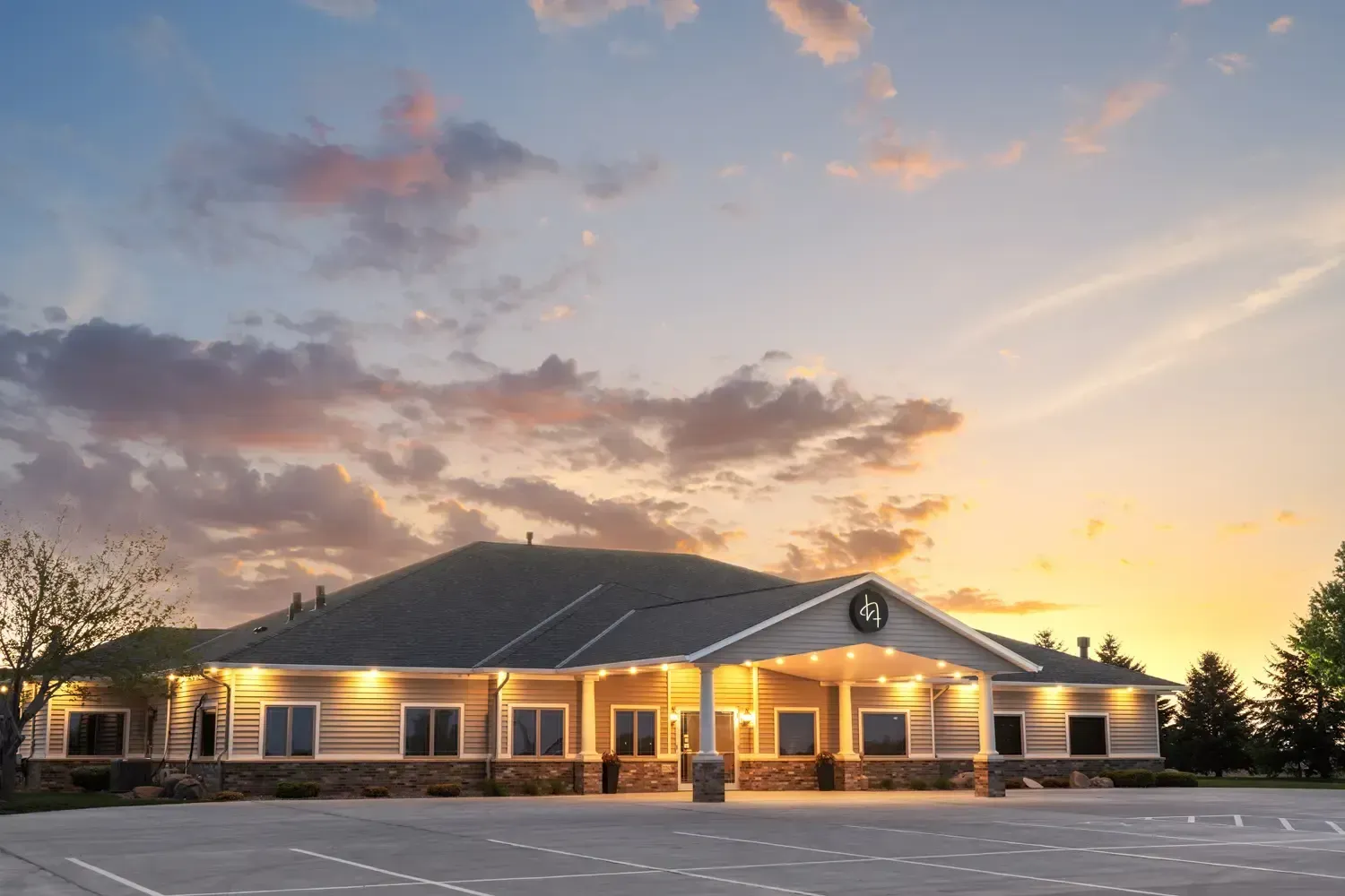 Single-story brick building at sunset with lit windows and a dark roof under a colorful sky