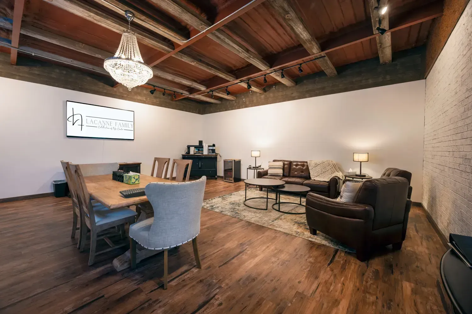 Rustic living room with exposed wood beams, long dining table, chairs, sofa, armchair, and wall-mounted TV
