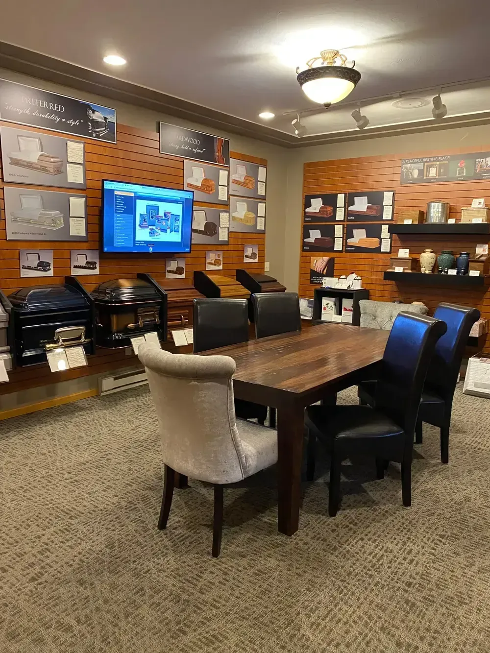 Conference room with a long wooden table, mixed chairs, wall-mounted TV, and shelves along wood-paneled walls.