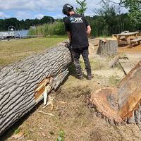 A firefighter in protective gear uses a chainsaw to cut a tree trunk outdoors, with a red crane truck in the background.