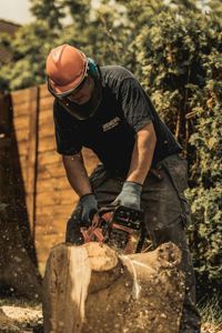 A person wearing safety gear, including a hard hat and face shield, uses a chainsaw to cut a wooden log outdoors.