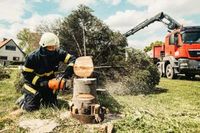 A firefighter in protective gear uses a chainsaw to cut a tree trunk outdoors, with a red crane truck in the background.