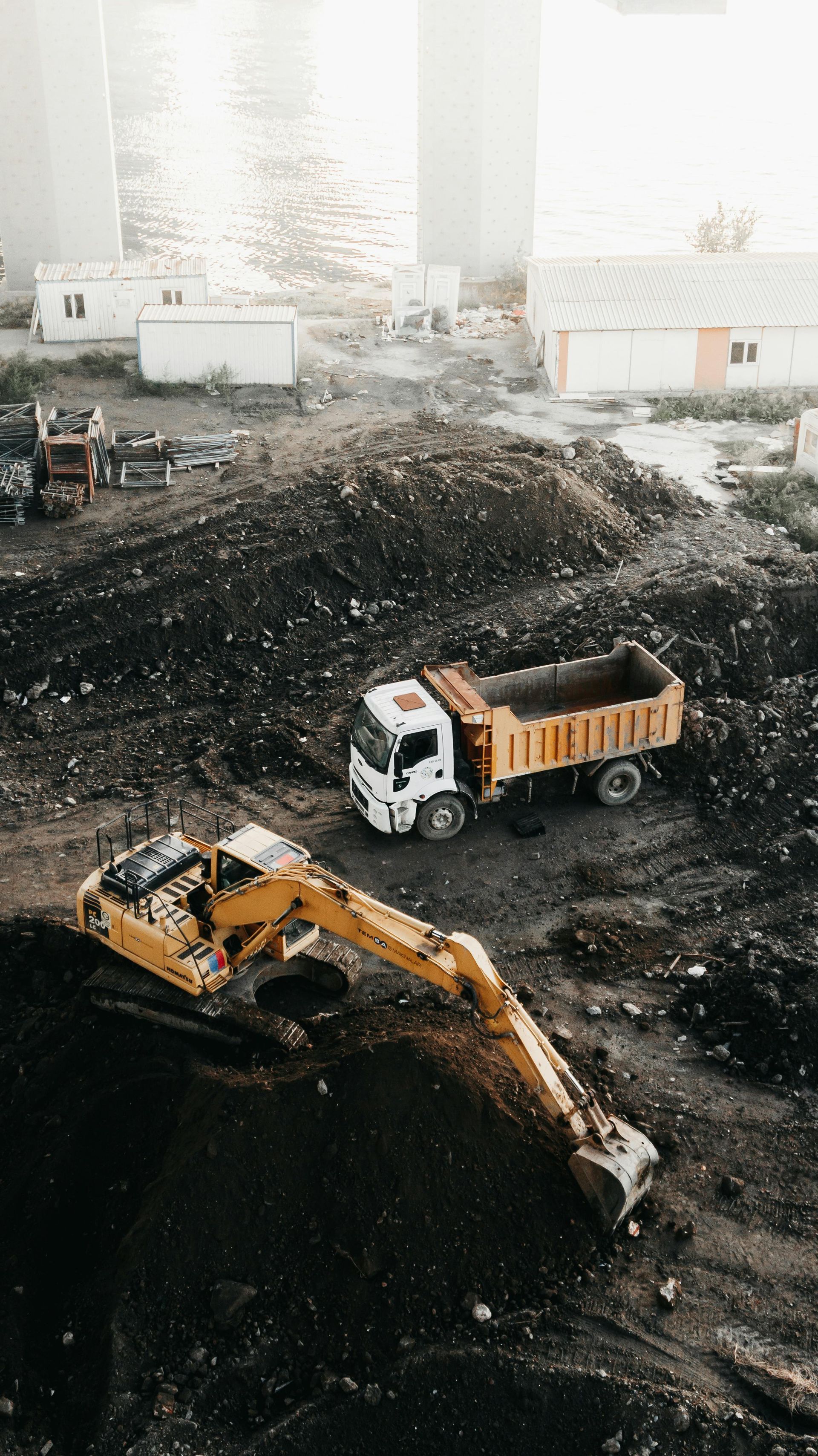 A yellow excavator works near a white dump truck on a pile of dark earth at a construction site.