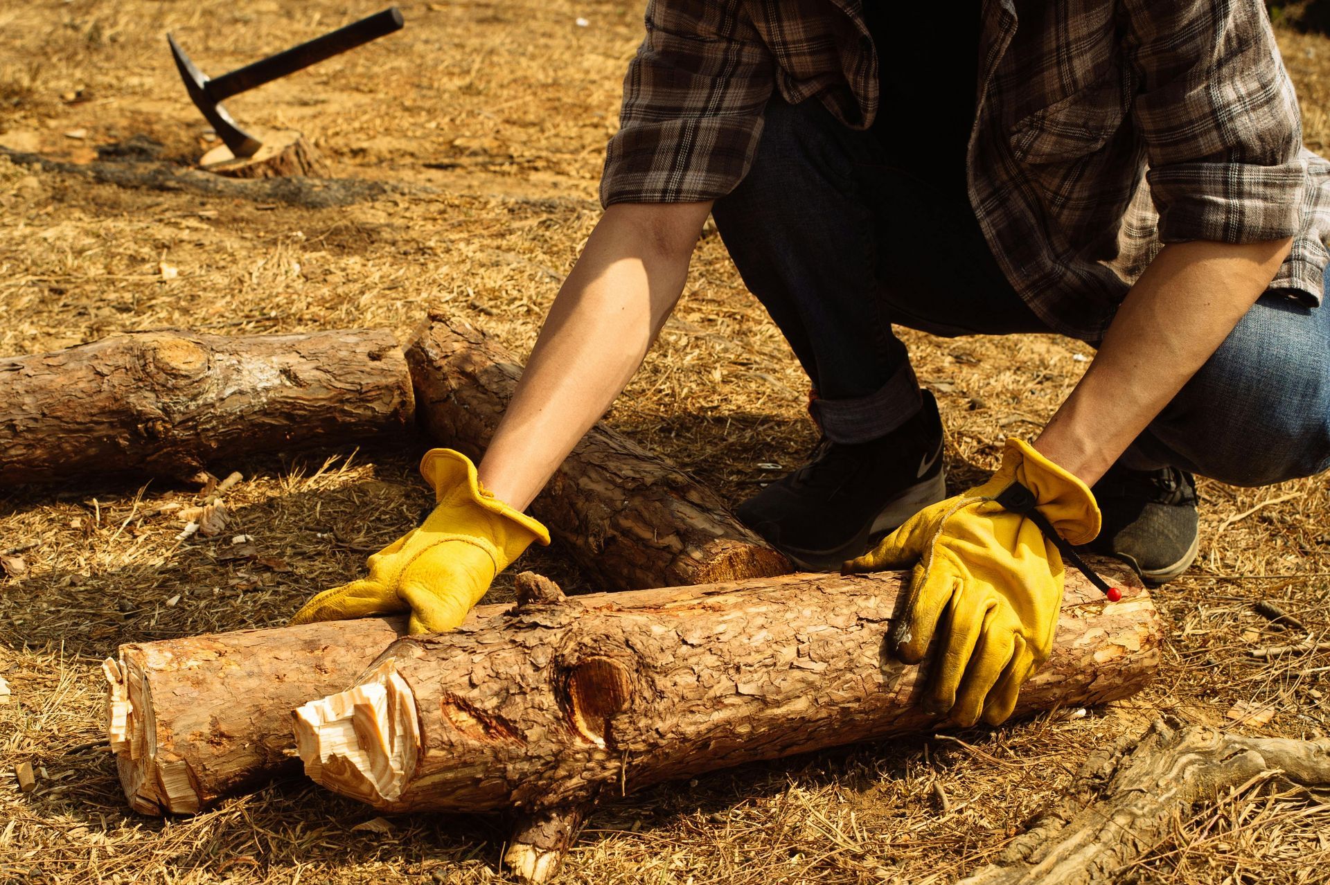 A person in a plaid shirt and yellow work gloves crouches on wood chips, positioning a large log near a tool.