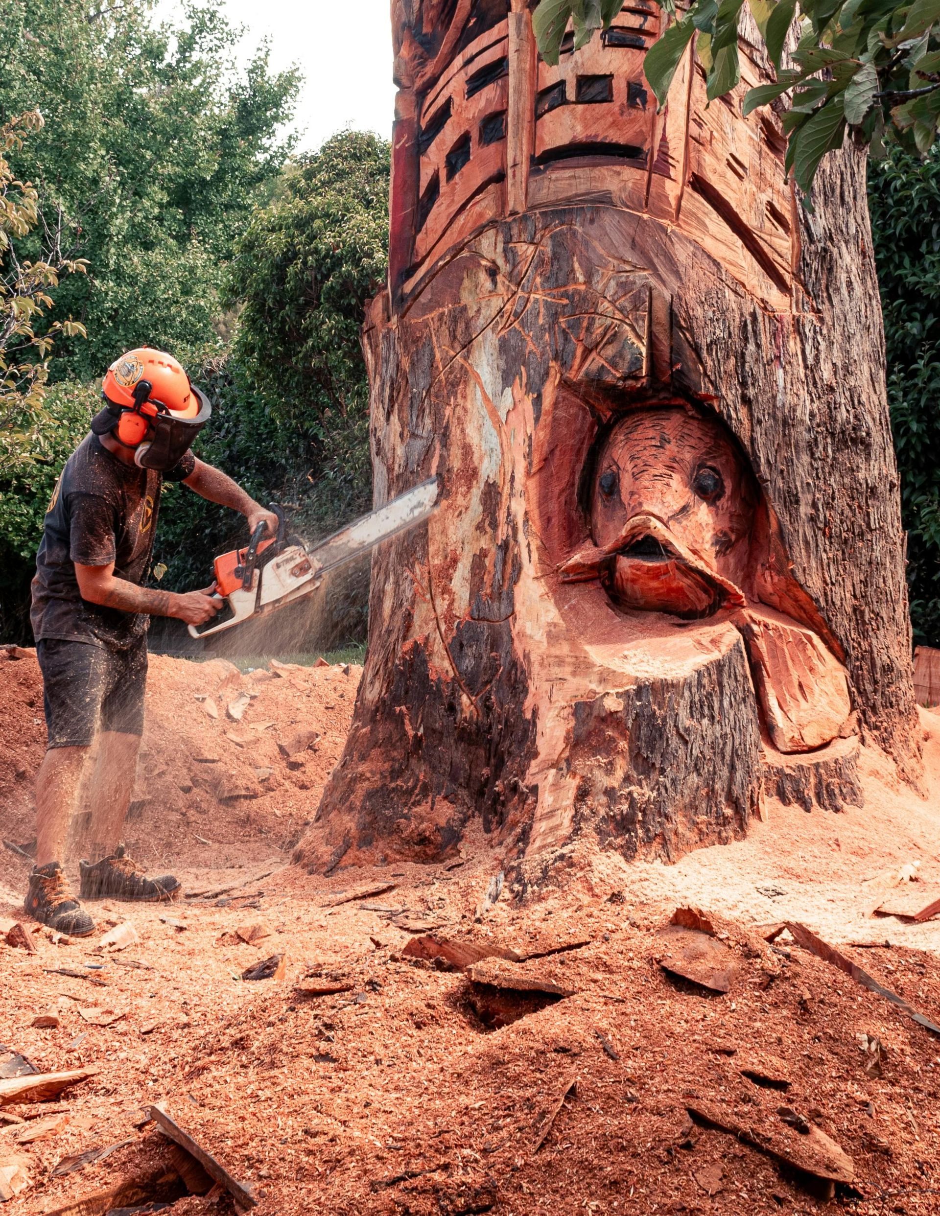 A person using a chainsaw to carve a fish sculpture out of a large tree trunk in an outdoor setting.