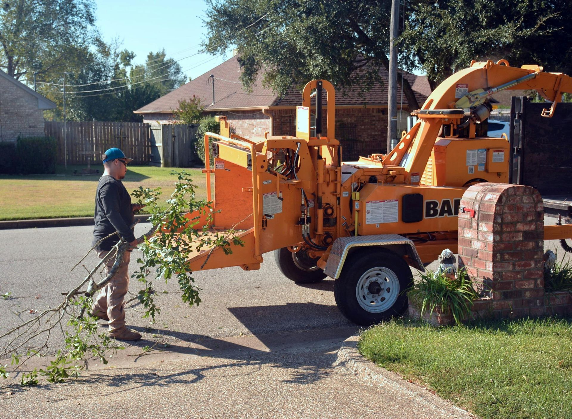 A worker in a blue cap feeds tree branches into a large yellow wood chipper on a suburban street.