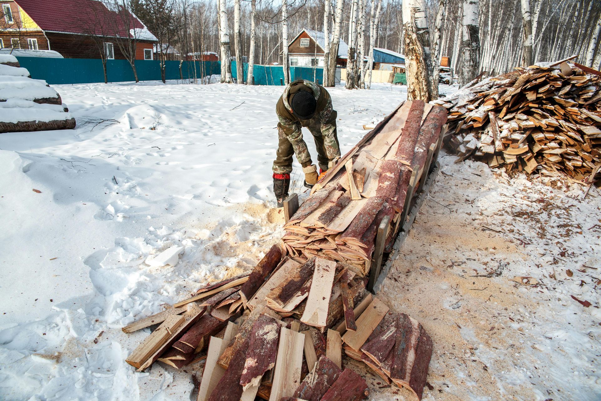 A person in camouflage gear uses a chainsaw to cut a log on a snow-covered ground near a pile of firewood.