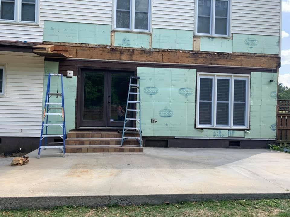 House exterior under construction, with exposed wall, insulation, doors, windows, ladders on concrete patio.
