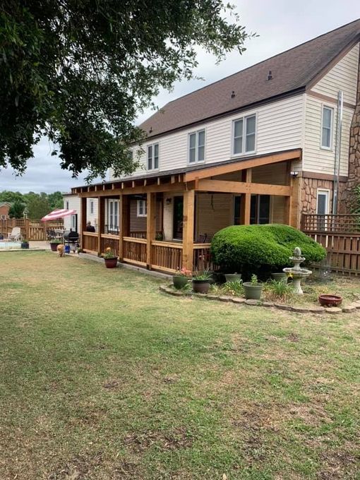 Large house with a wooden porch. Brown siding and trim. Green grass, cloudy sky.