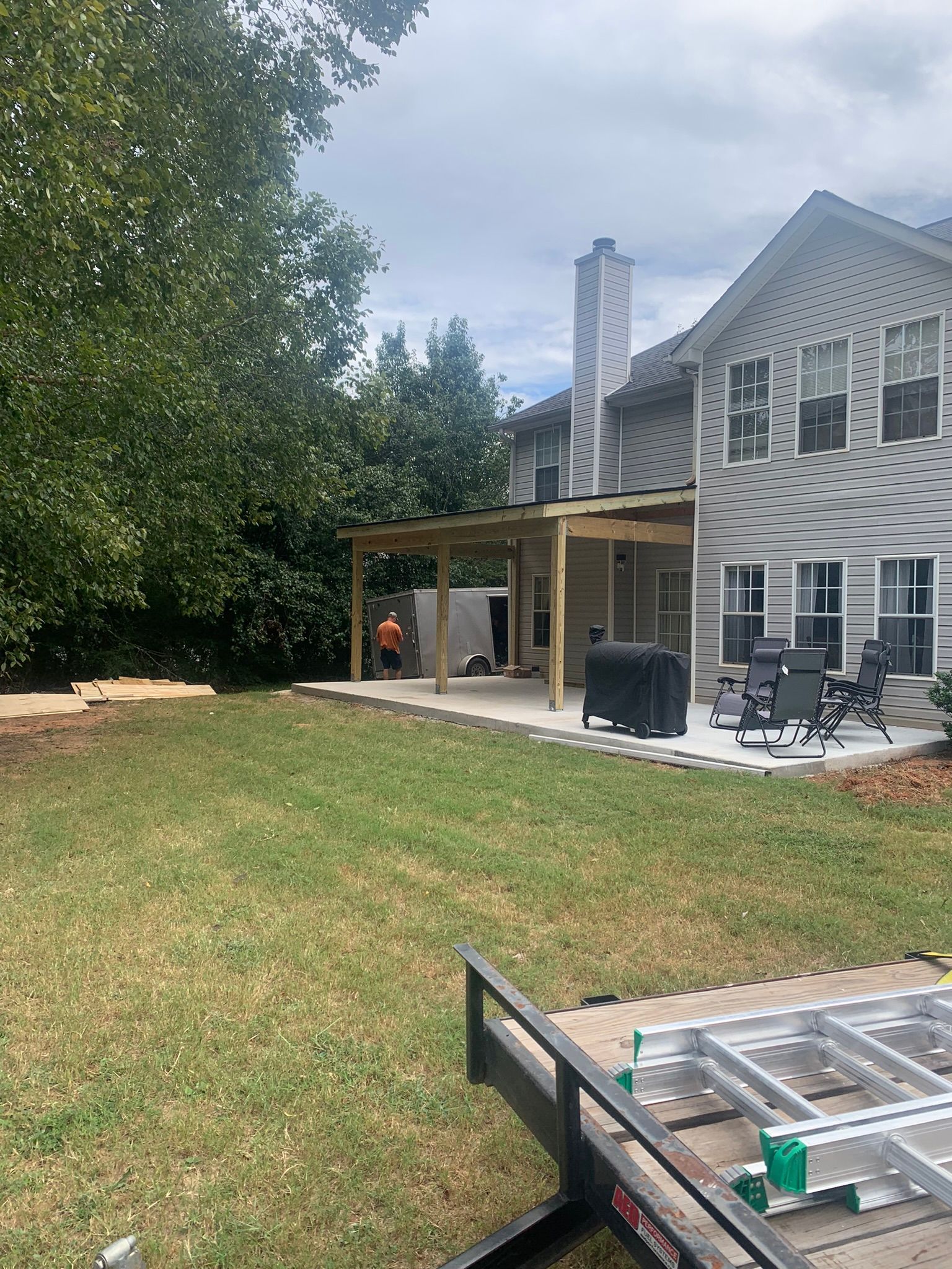 Backyard with new covered patio under construction; worker on site, trailer, and green grass.