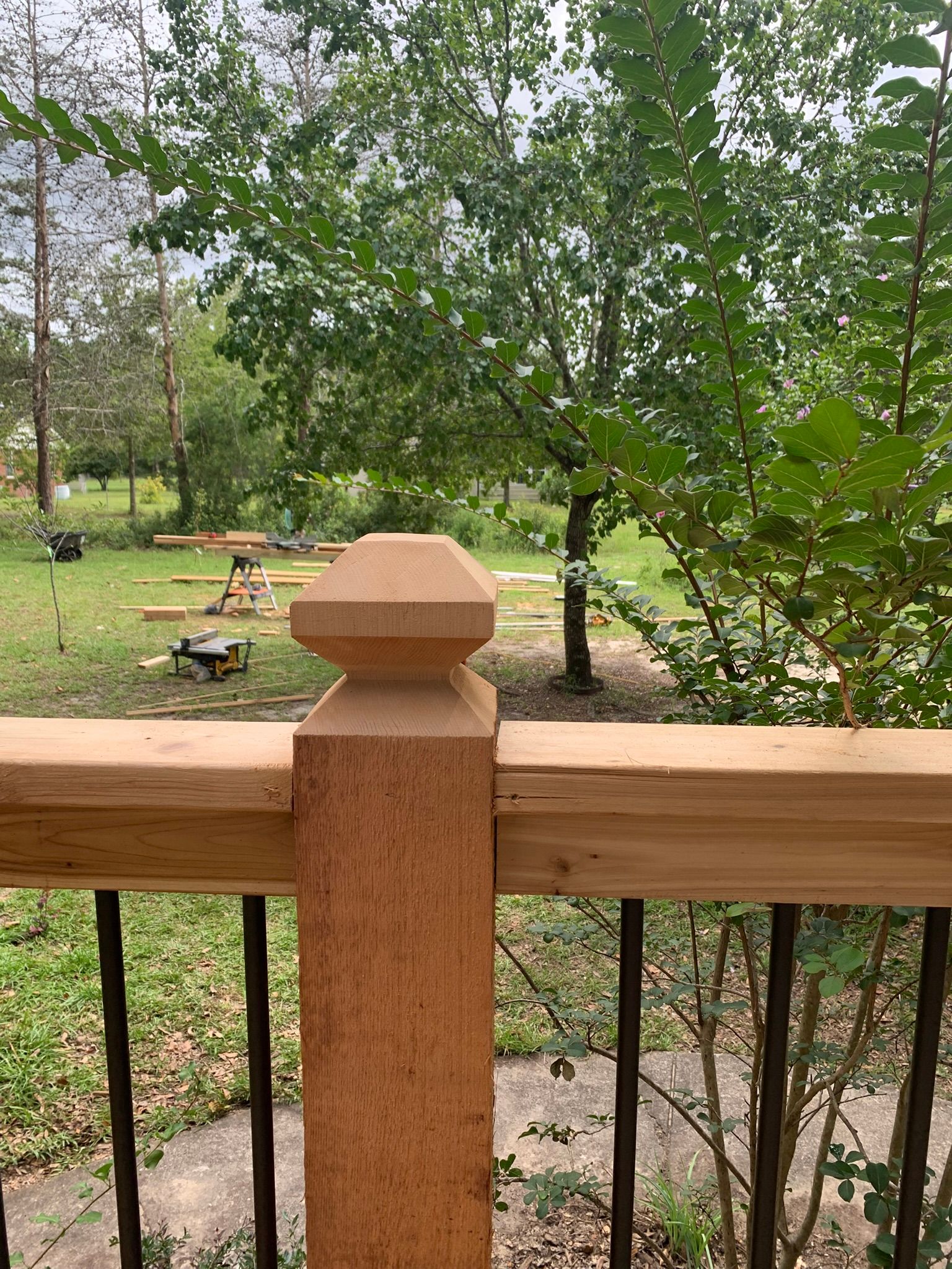 Wooden deck railing with a decorative post overlooking a backyard with trees and construction in progress.