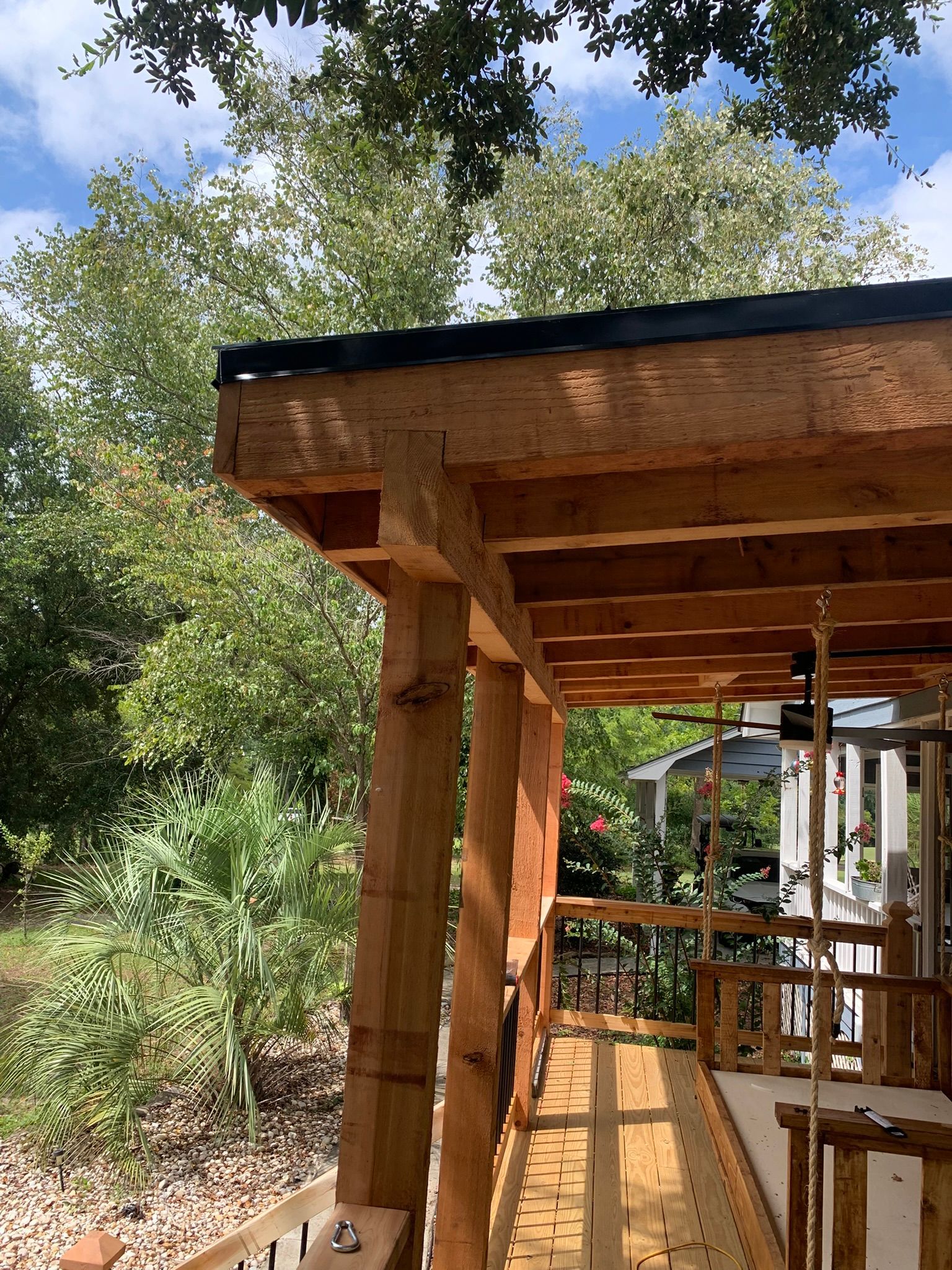 Wooden porch with exposed beams and supporting posts, surrounded by lush greenery and a blue sky.
