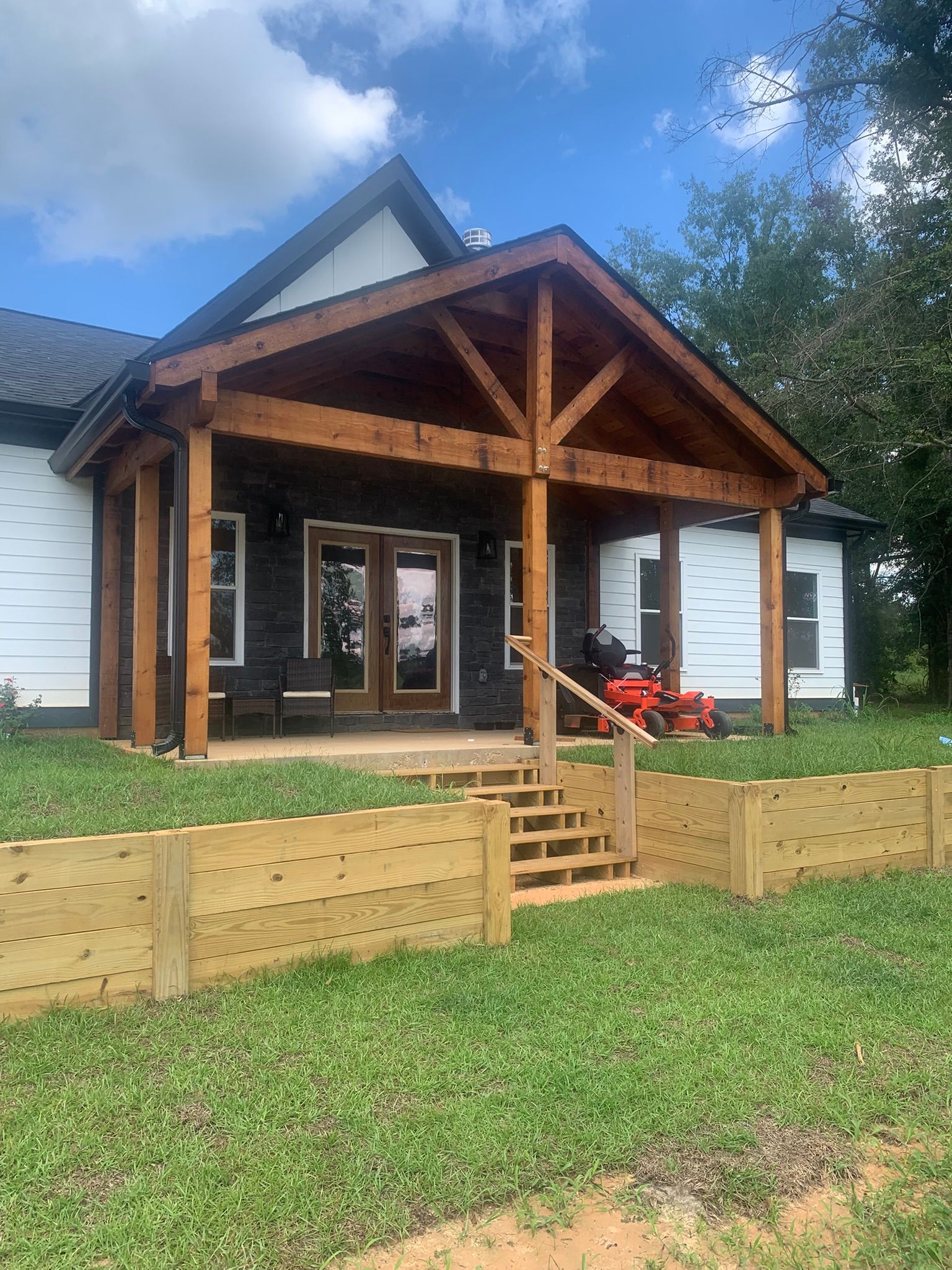 A wooden porch with a dark roof and black siding. Wooden retaining walls and lawn in the foreground.