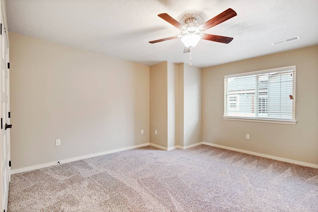 Empty bedroom with beige walls, carpet, window, and ceiling fan.
