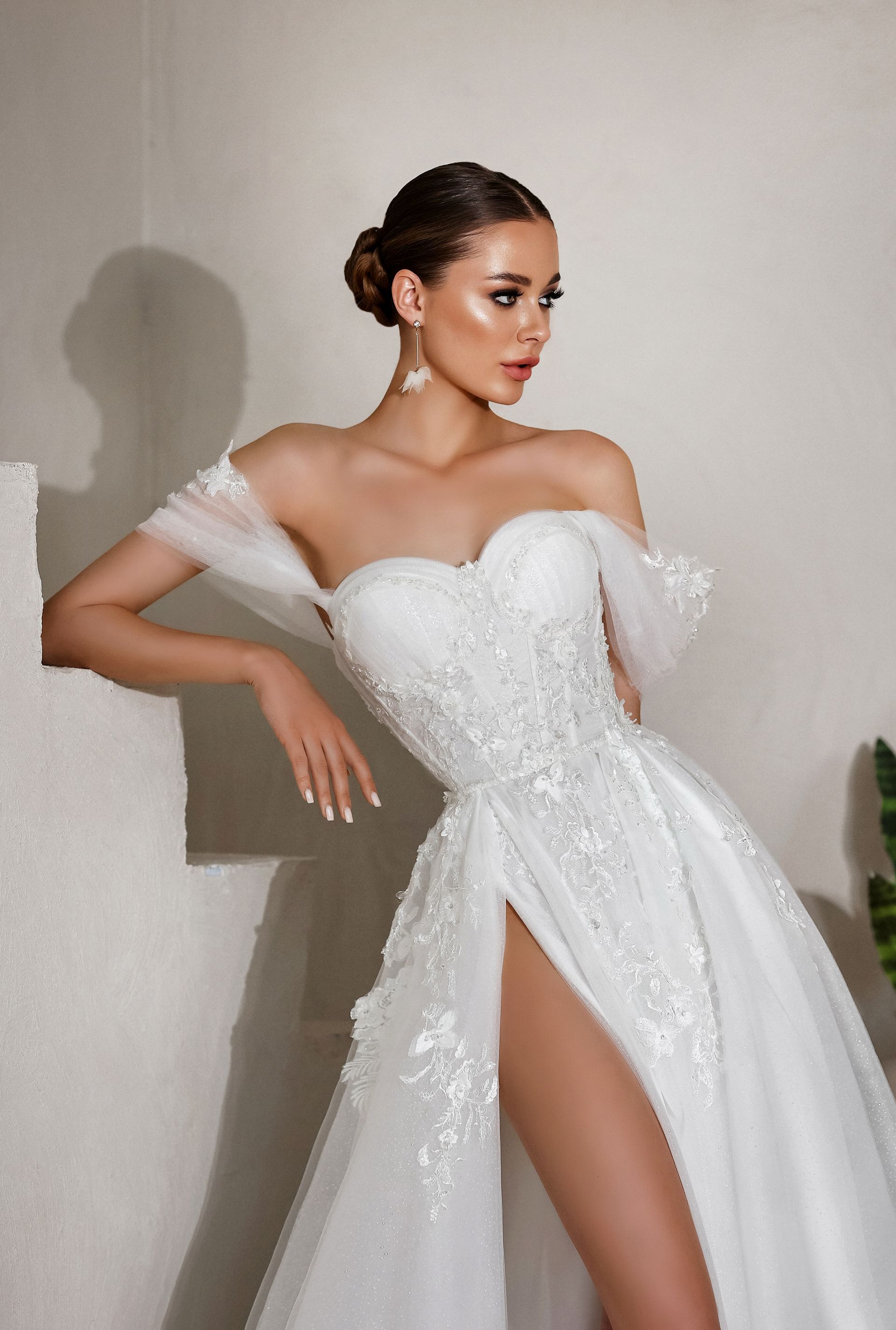 Woman in a white off-shoulder gown posing against a plain wall, with a high slit and sparkling details.
