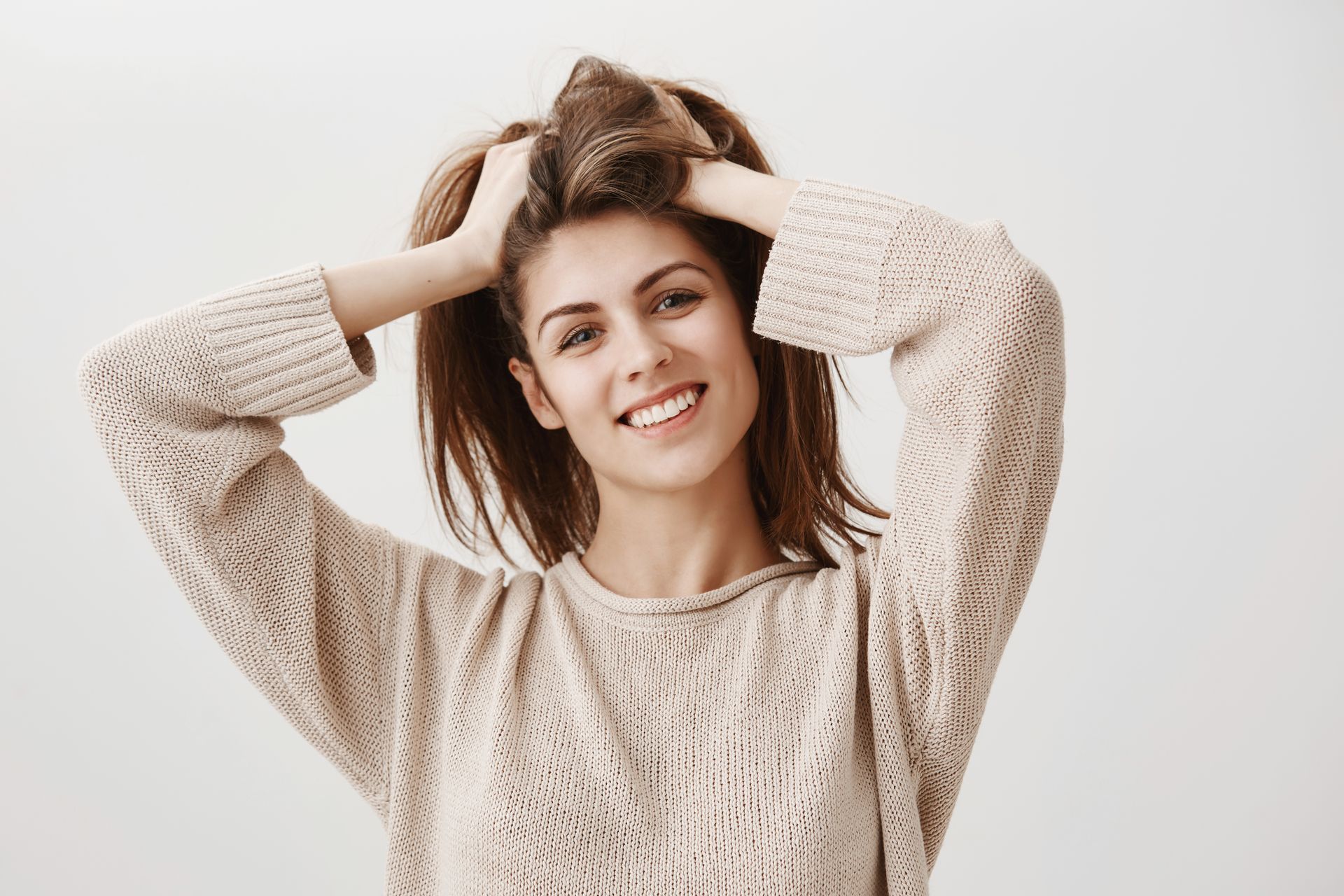 Person smiling with hands in hair, wearing a beige sweater against a light background