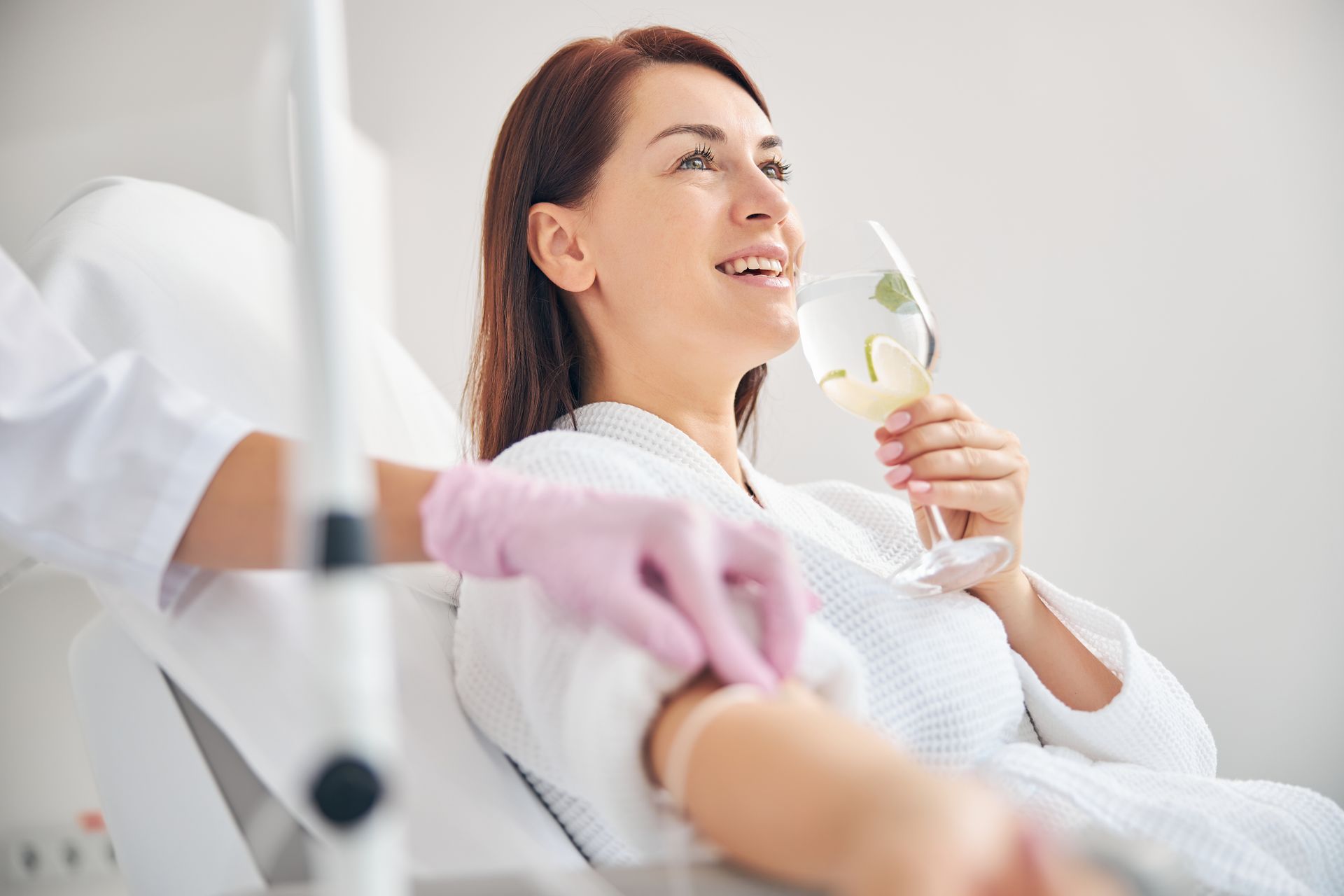 Woman reclining in a clinic chair, smiling while holding an IV drip bag, with a nurse nearby