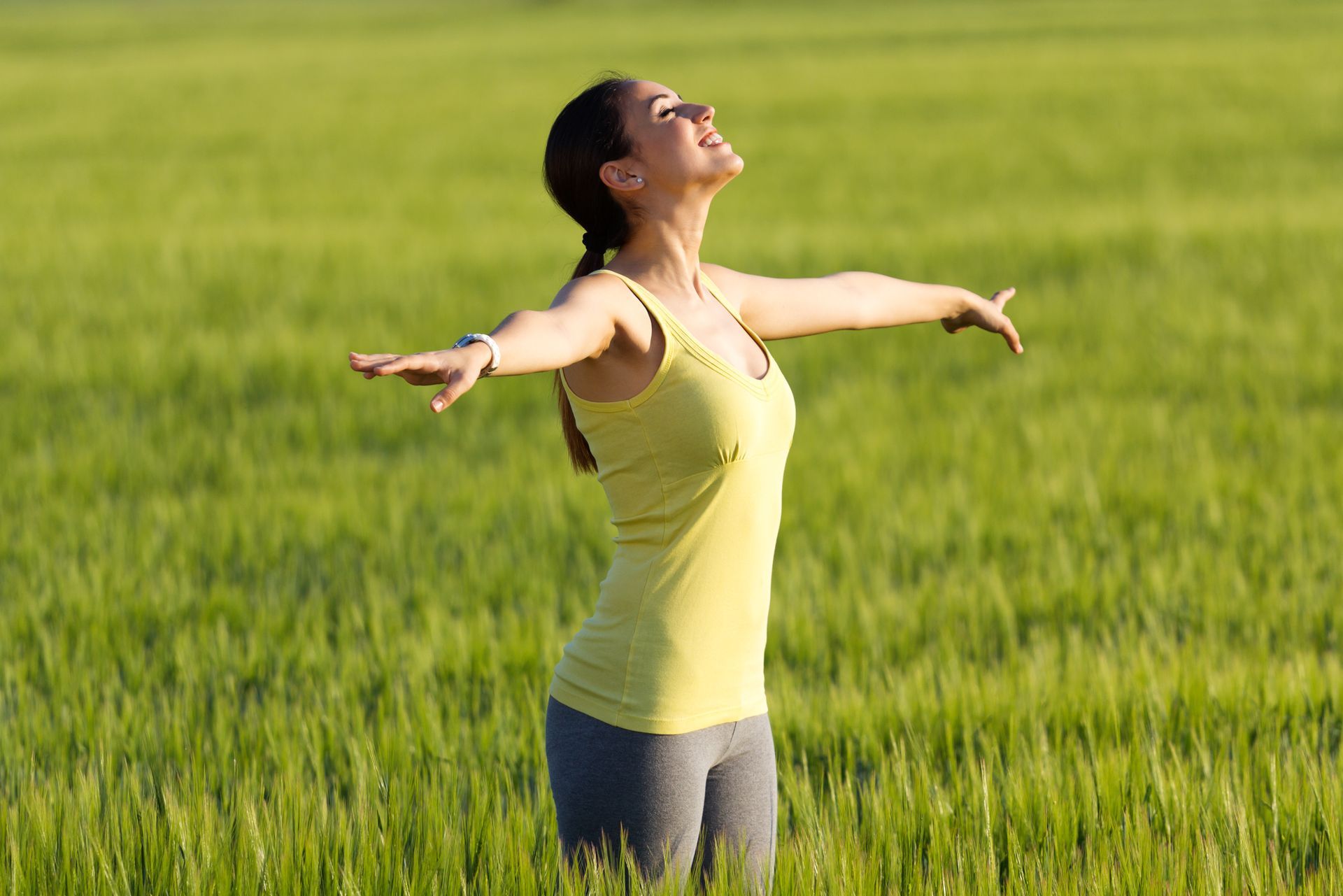 Woman in a yellow top standing in a green field with arms outstretched, looking up