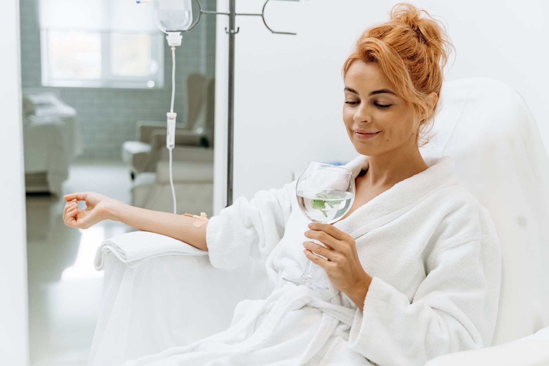 Woman in a white robe resting in a medical chair, holding a drink in a bright clinic room