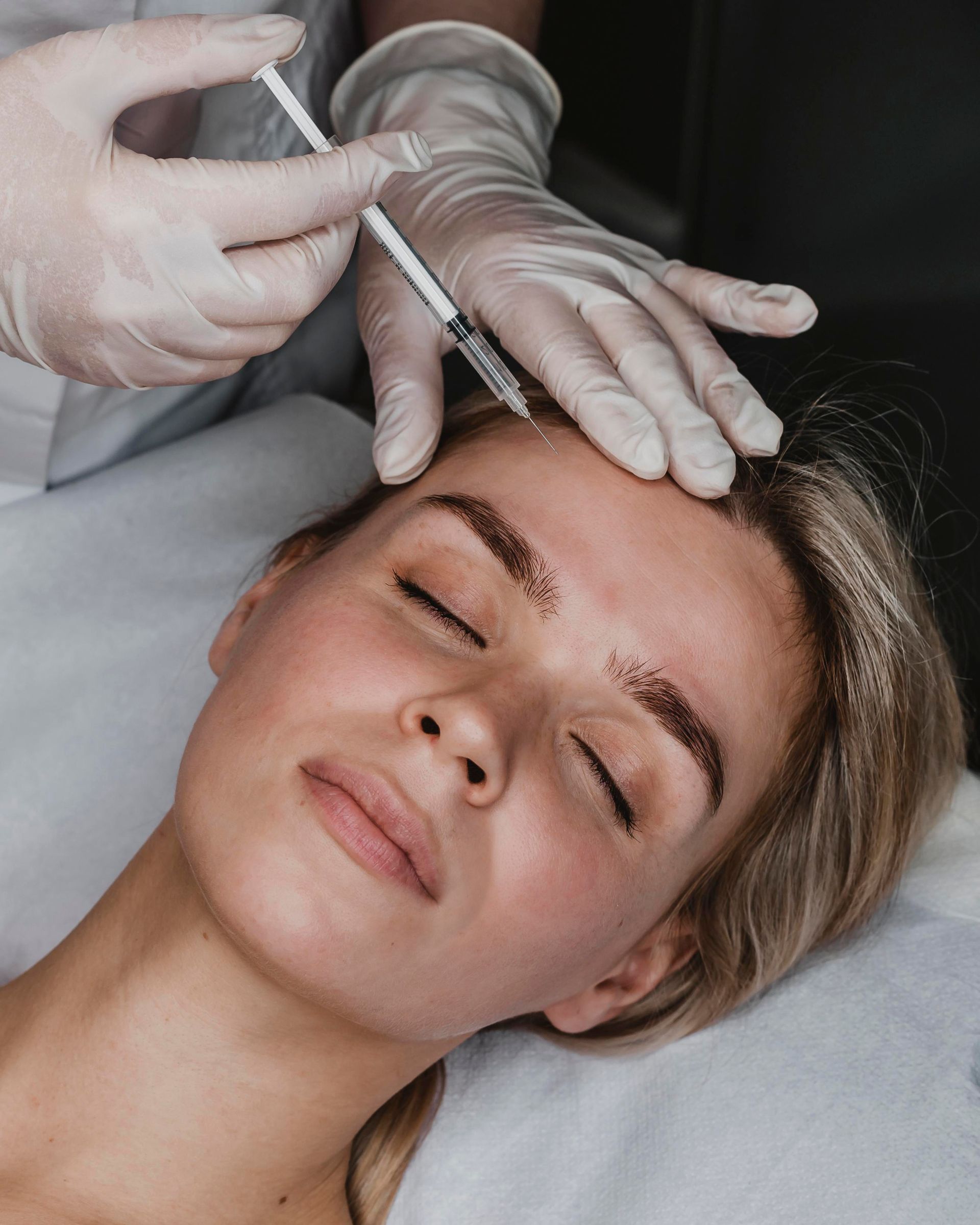 Gloved hands administer a facial injection to a reclining woman with eyes closed