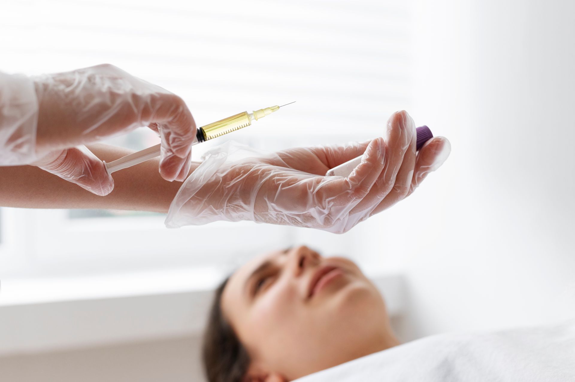 Gloved hands preparing a syringe above a reclining patient in a bright clinical setting