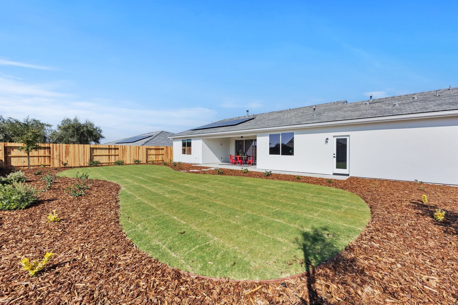 Backyard with grass, mulch border, wooden fence, and white house under a clear blue sky.