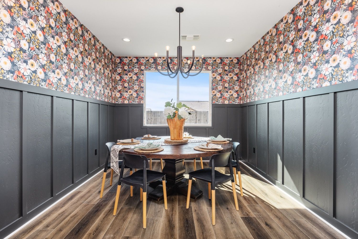 Dining room with floral wallpaper, dark wainscoting, round table set for dining, black chairs, and a chandelier.