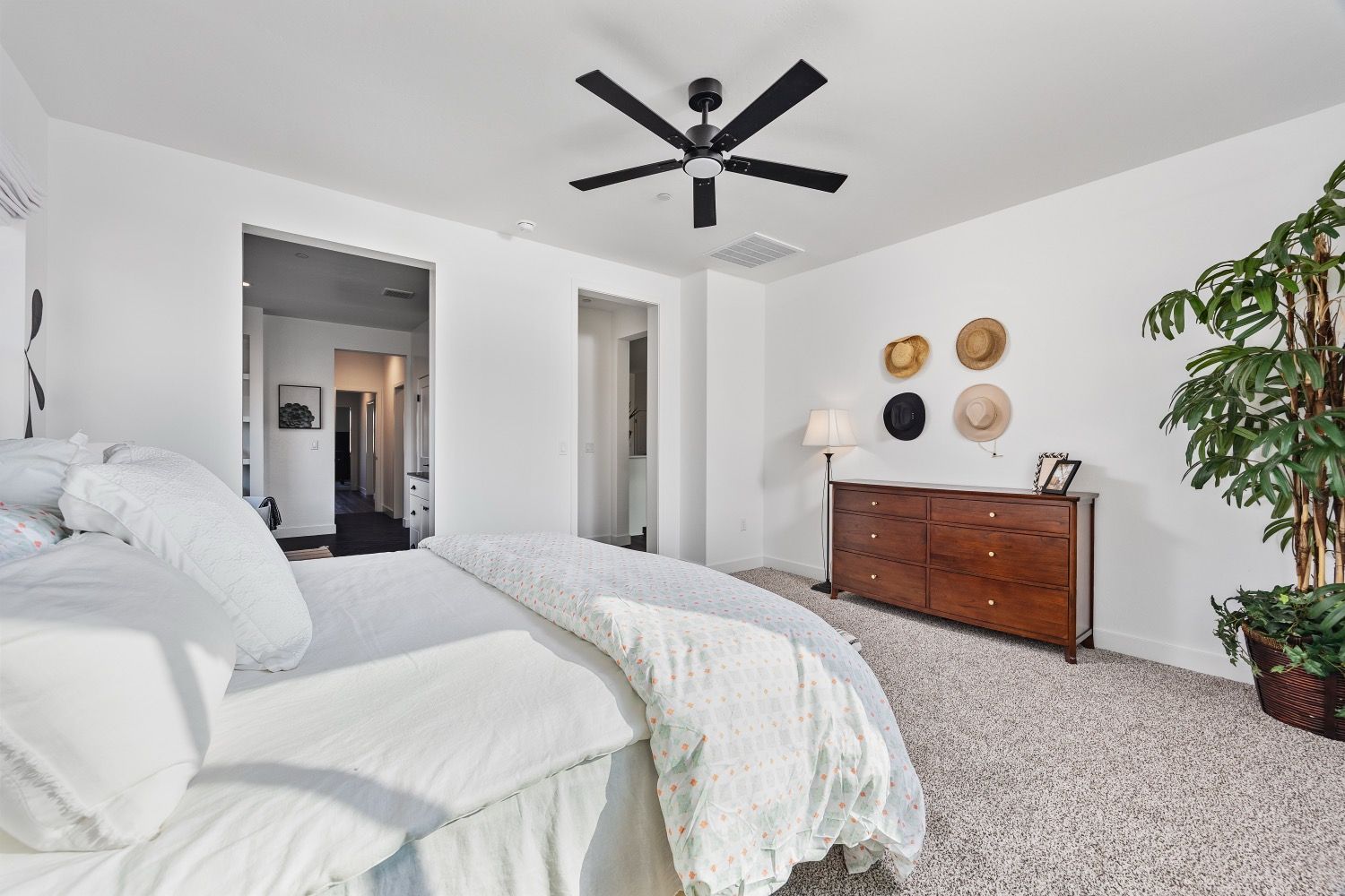 Bedroom with white bedding, wooden dresser, hats on wall, ceiling fan, and large potted plant.