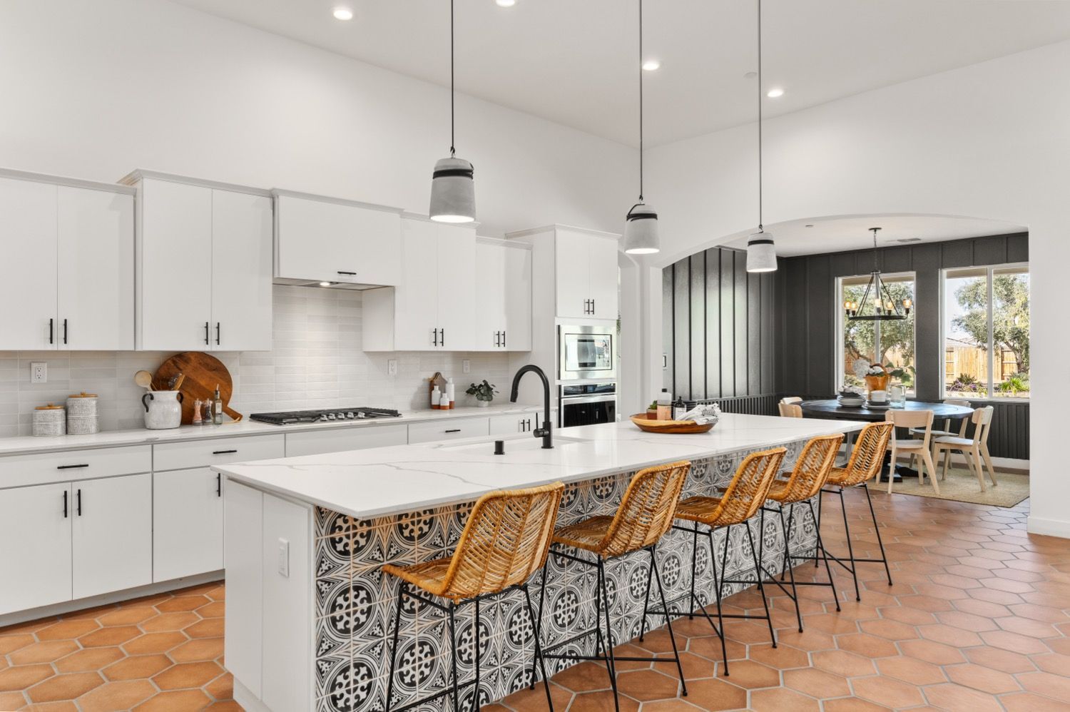 White kitchen with island, patterned tile, and woven bar stools. Dining area in the background with natural light.
