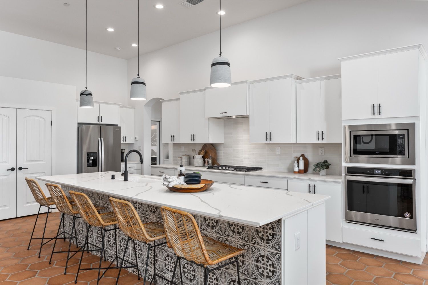 Modern white kitchen with island seating, stainless steel appliances, and terra-cotta flooring.