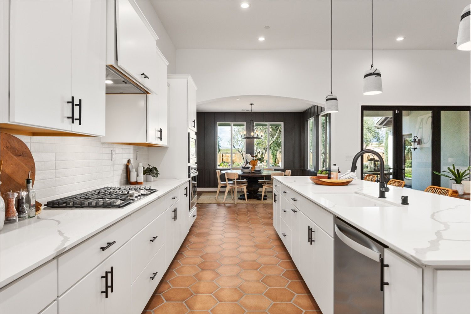 Modern white kitchen with long island and terracotta tile floor, leading to dining area.