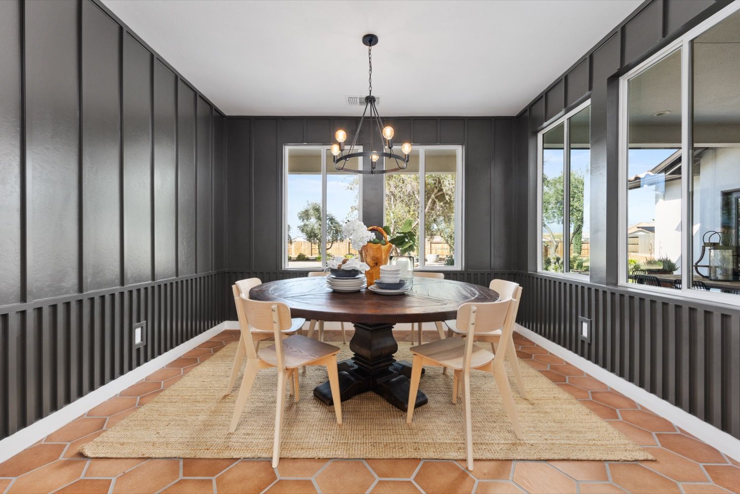 Dining room with dark gray paneled walls, round wooden table, chairs, and window overlooking a sunny outdoor scene.