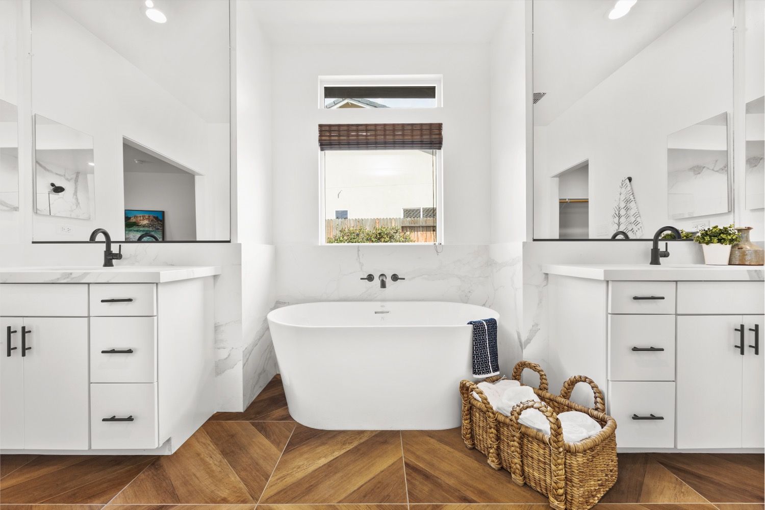 White bathroom with dual vanities, freestanding tub, and herringbone wood floor.