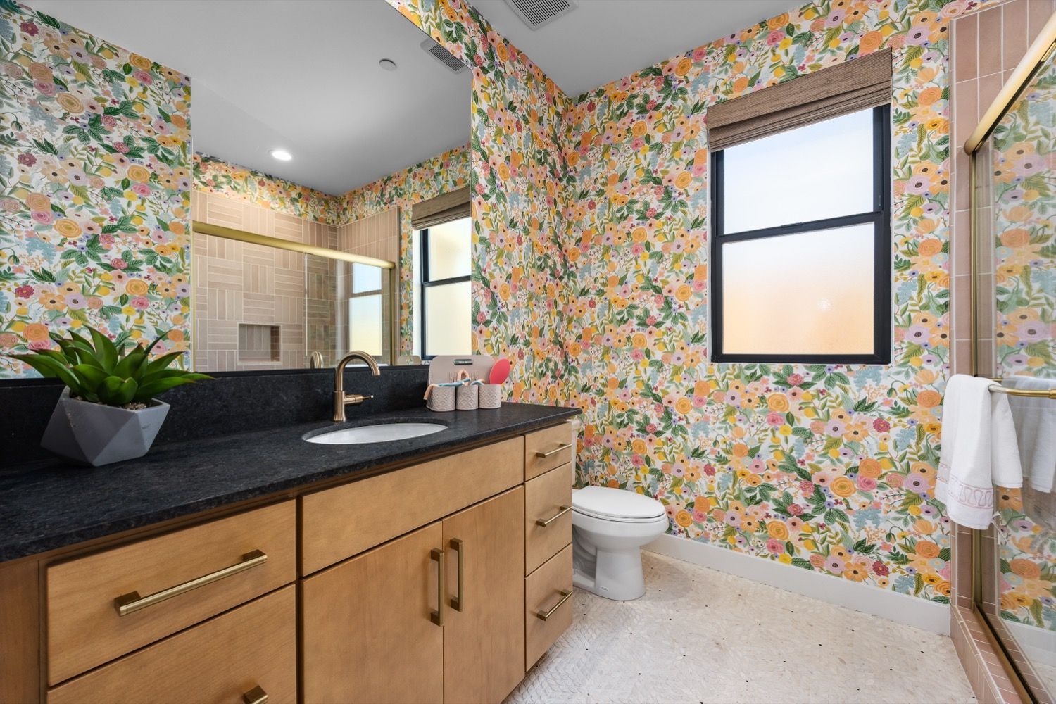 Bathroom with floral wallpaper, wood vanity, black countertop, white toilet, and window.