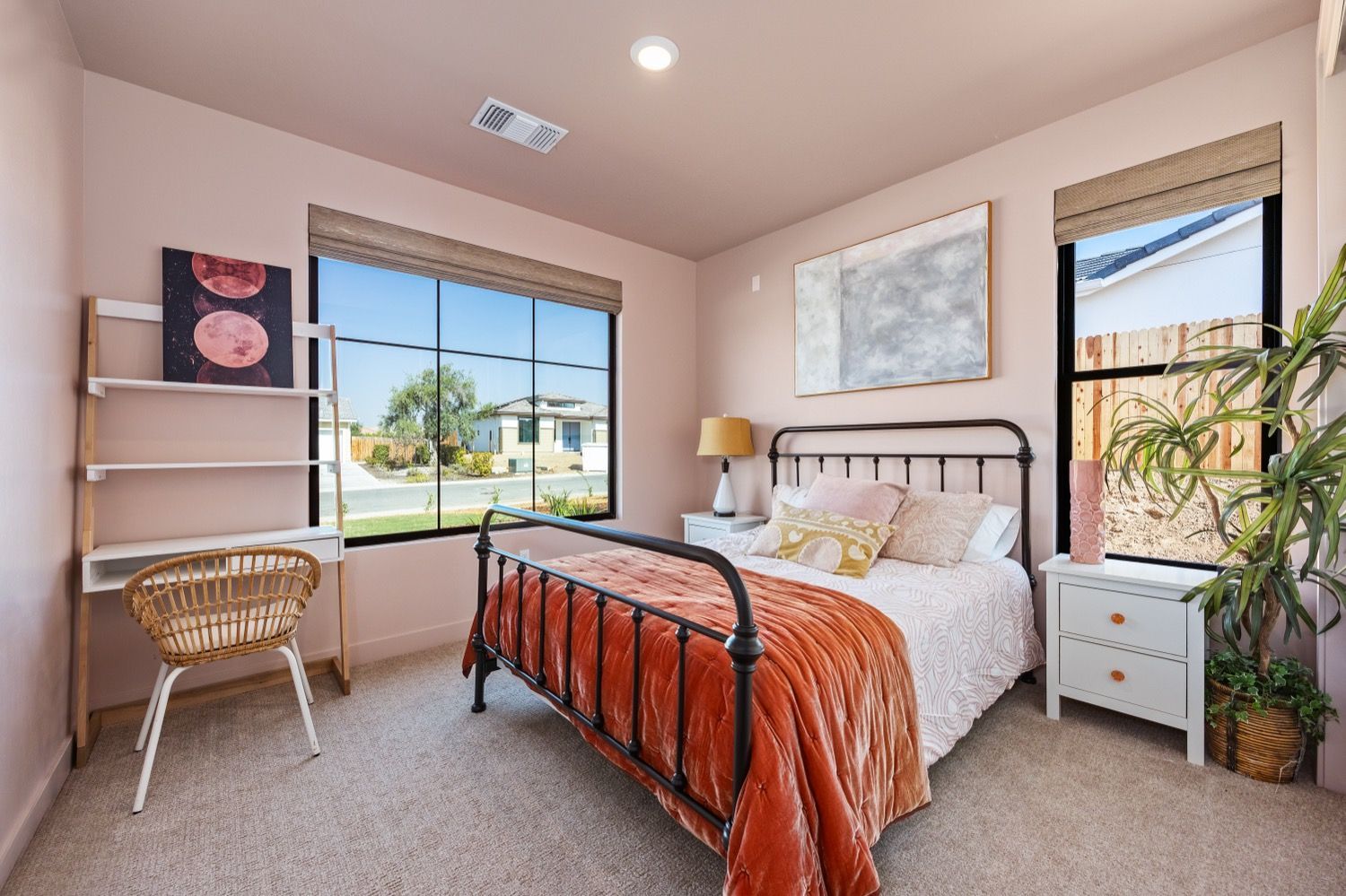 Bedroom with rose-colored walls, metal bed frame, large windows, desk, and decorative accents.